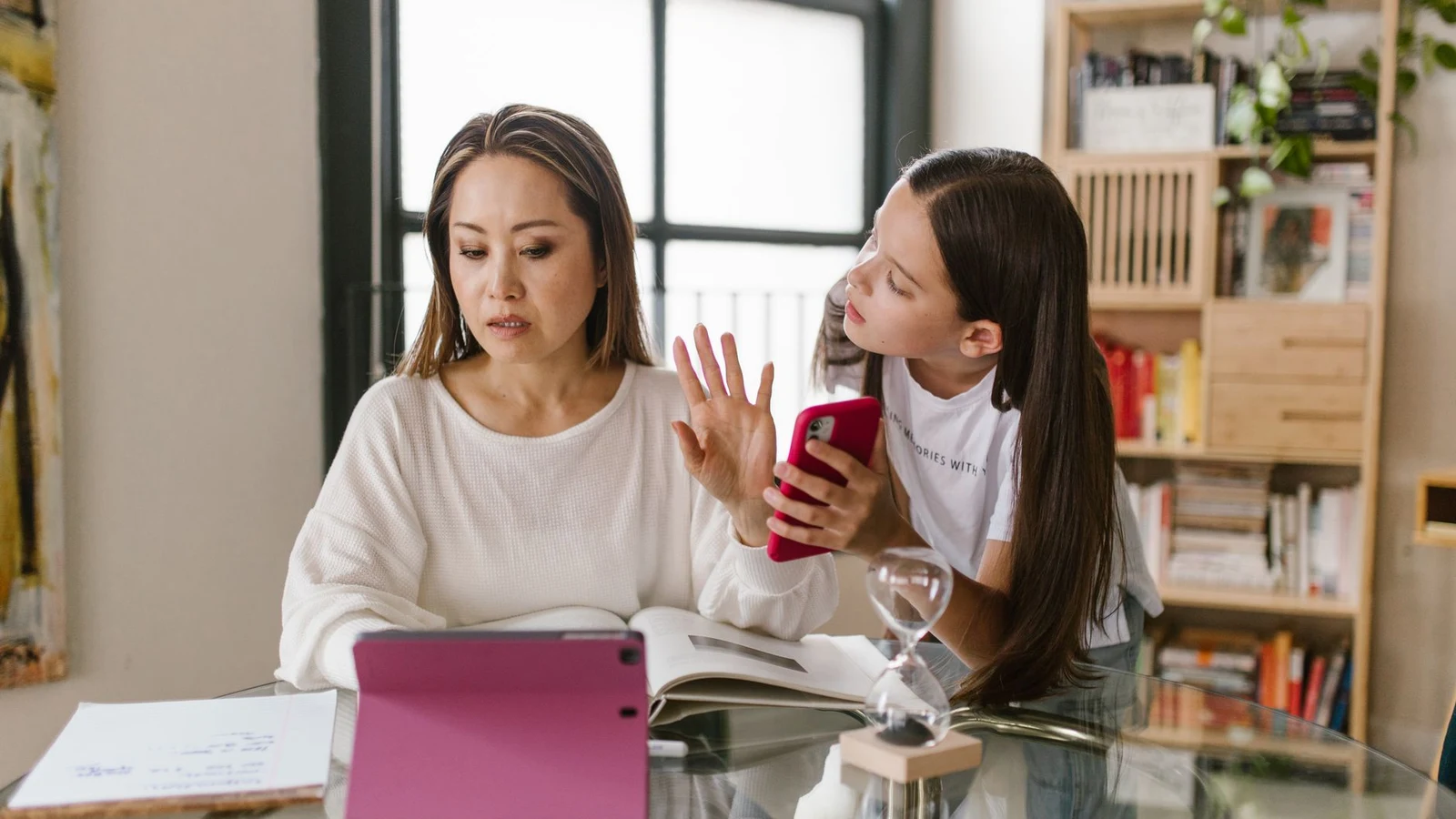 A mother and her tween daughter negotiating over a phone at the kitchen table, with an hourglass sitting between them