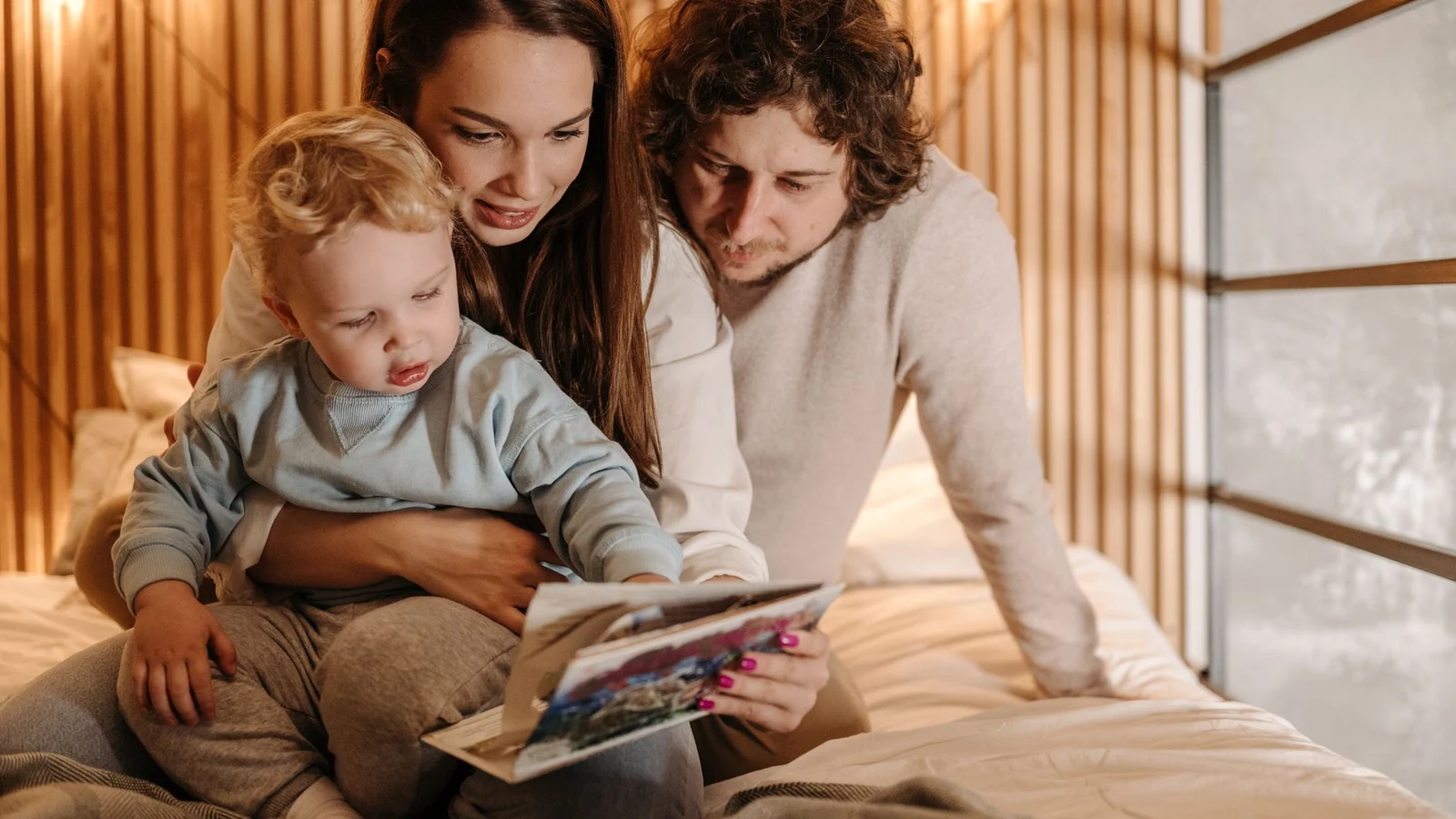 A toddler sitting between both parents on a bed, all three absorbed in an open book together in warm evening light