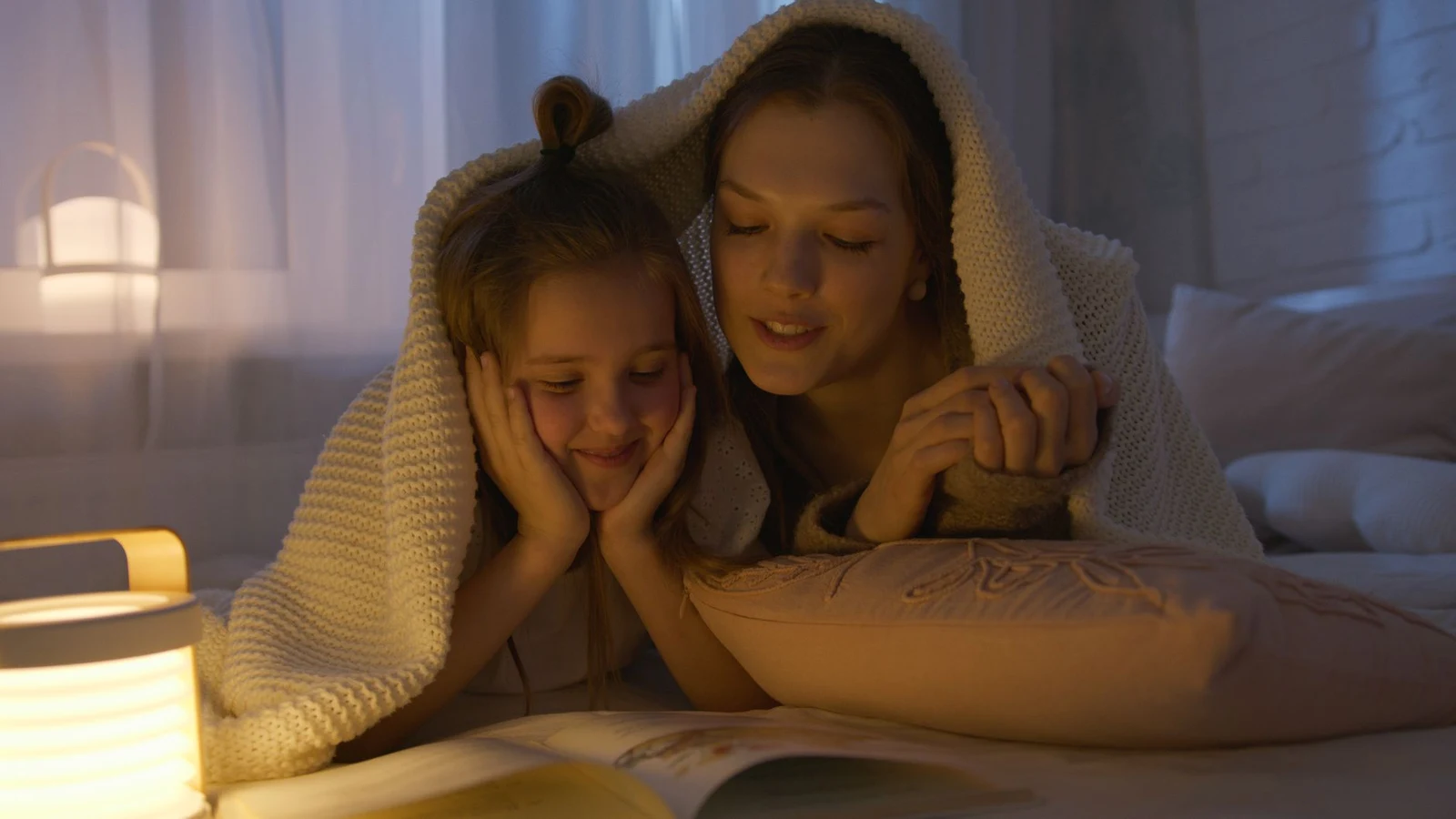 Mother and daughter reading together under a blanket by warm lamp light during a calm bedtime wind-down